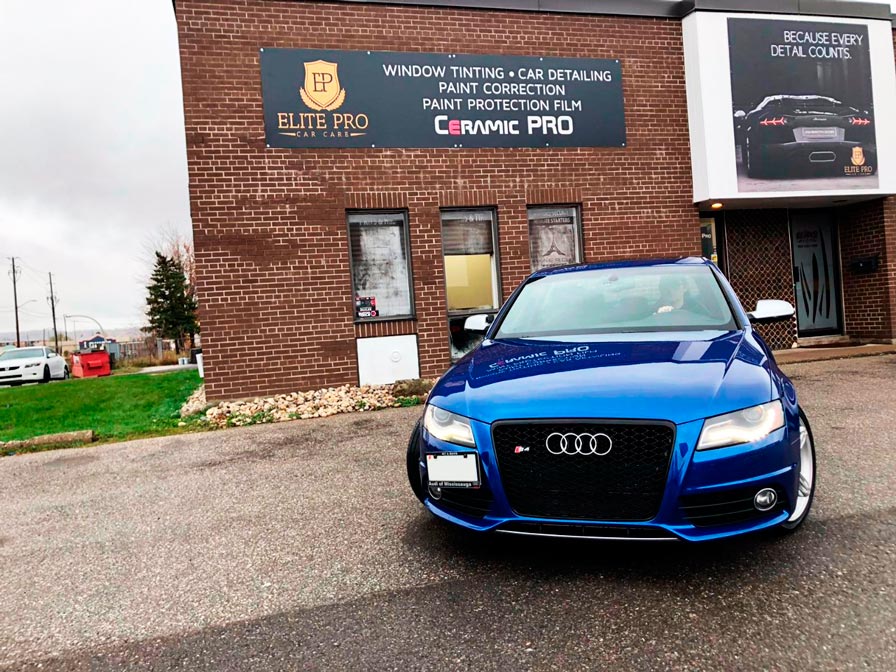 Blue car parked in front of a brick building with Elite Pro Car Care signage, offering window tinting, car detailing, paint correction, paint protection film, and ceramic coating services